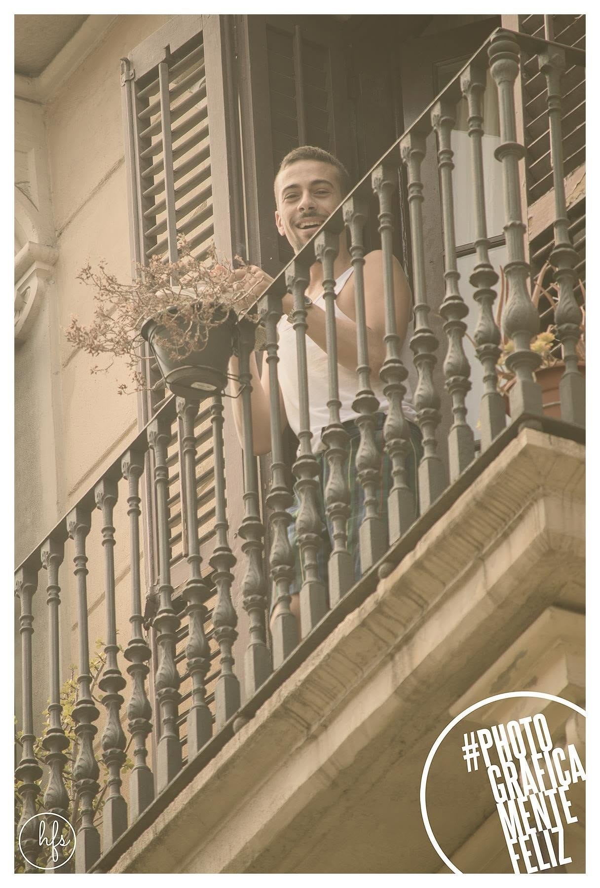 Hombre joven sonriente asomado a un balcón de hierro forjado con persianas de madera, sosteniendo una maceta con planta seca, visto desde abajo con la fachada de un edificio antiguo de…