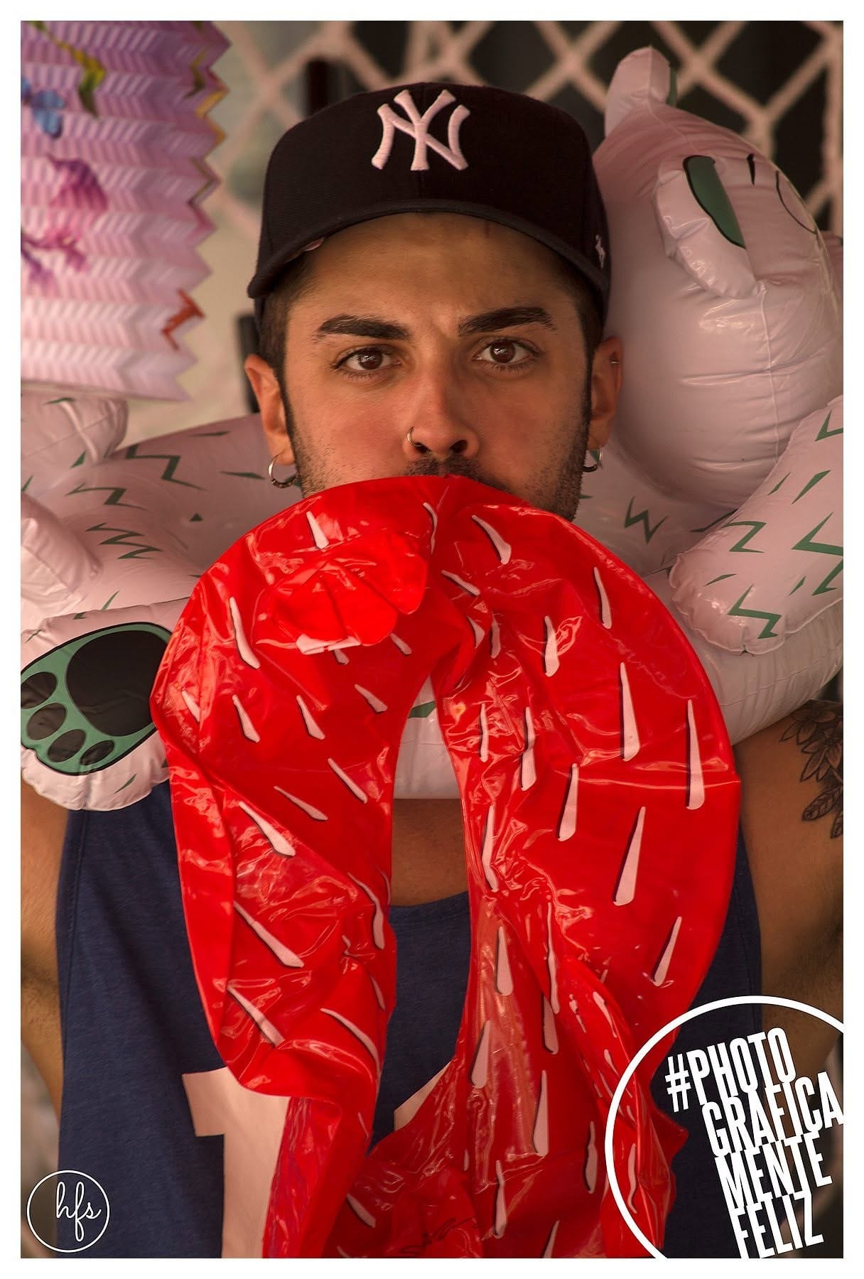 Retrato de un joven con gorra de los Yankees, piercing en el septum, sosteniendo frente a la boca una máscara o boa inflable roja con detalles blancos que simulan sangre, rodeado de otros…