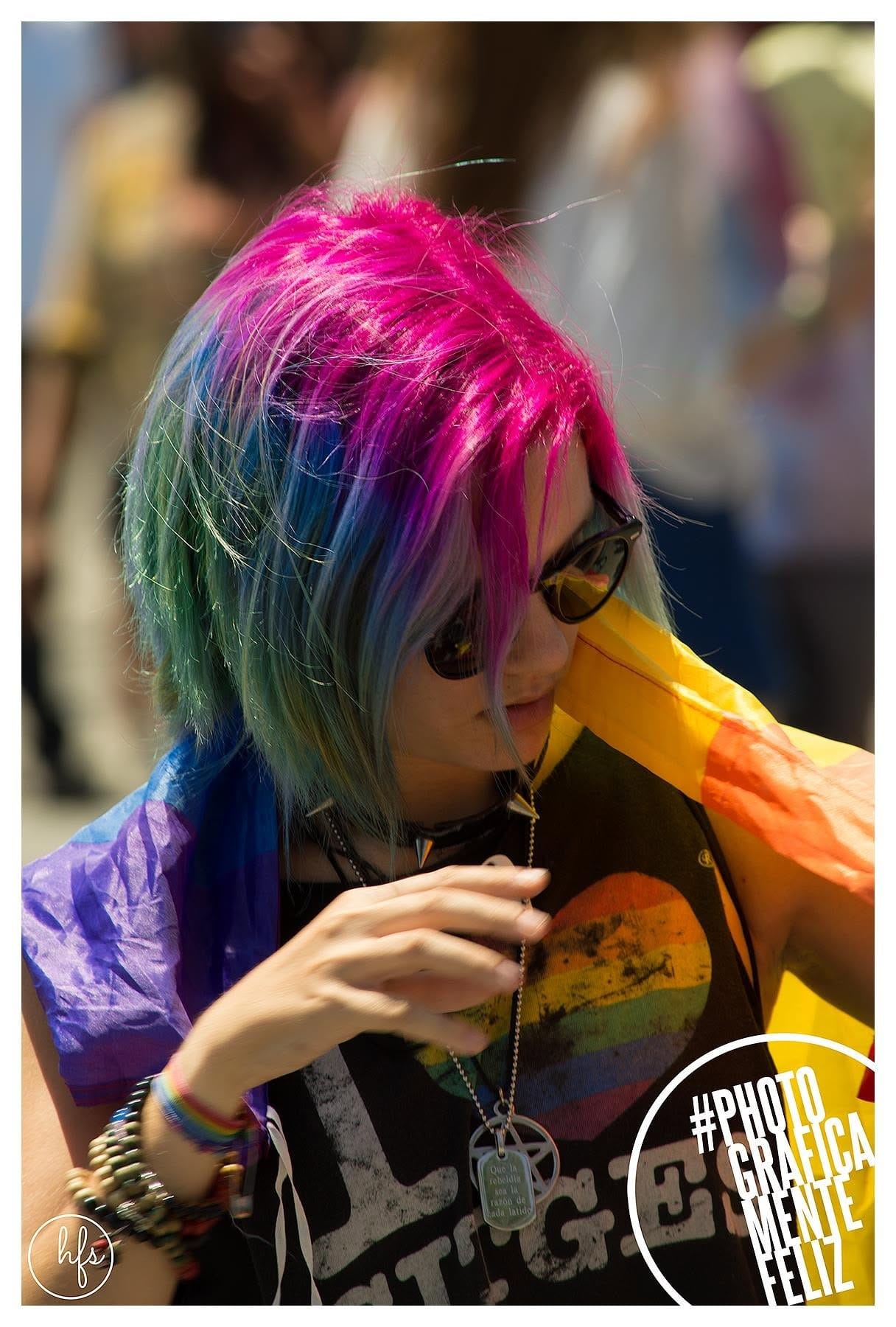 Retrato de una persona con el pelo pintado en tonos rosa, azul y verde, anteojos de sol oscuros y una bandera arcoíris sobre los hombros.