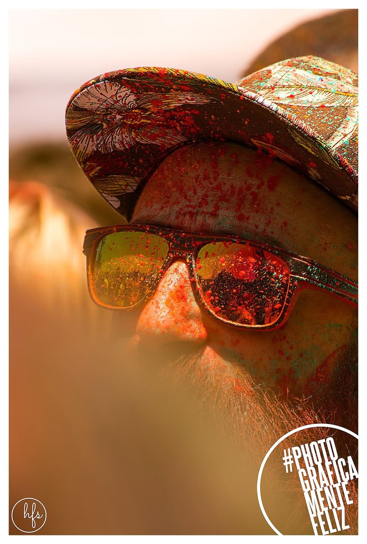 Retrato de un hombre con barba pelirroja cubierto de polvo naranja, con un sombrero de ala ancha estampado, gafas de sol reflectantes, visto en un contraluz cálido que difumina el fondo.