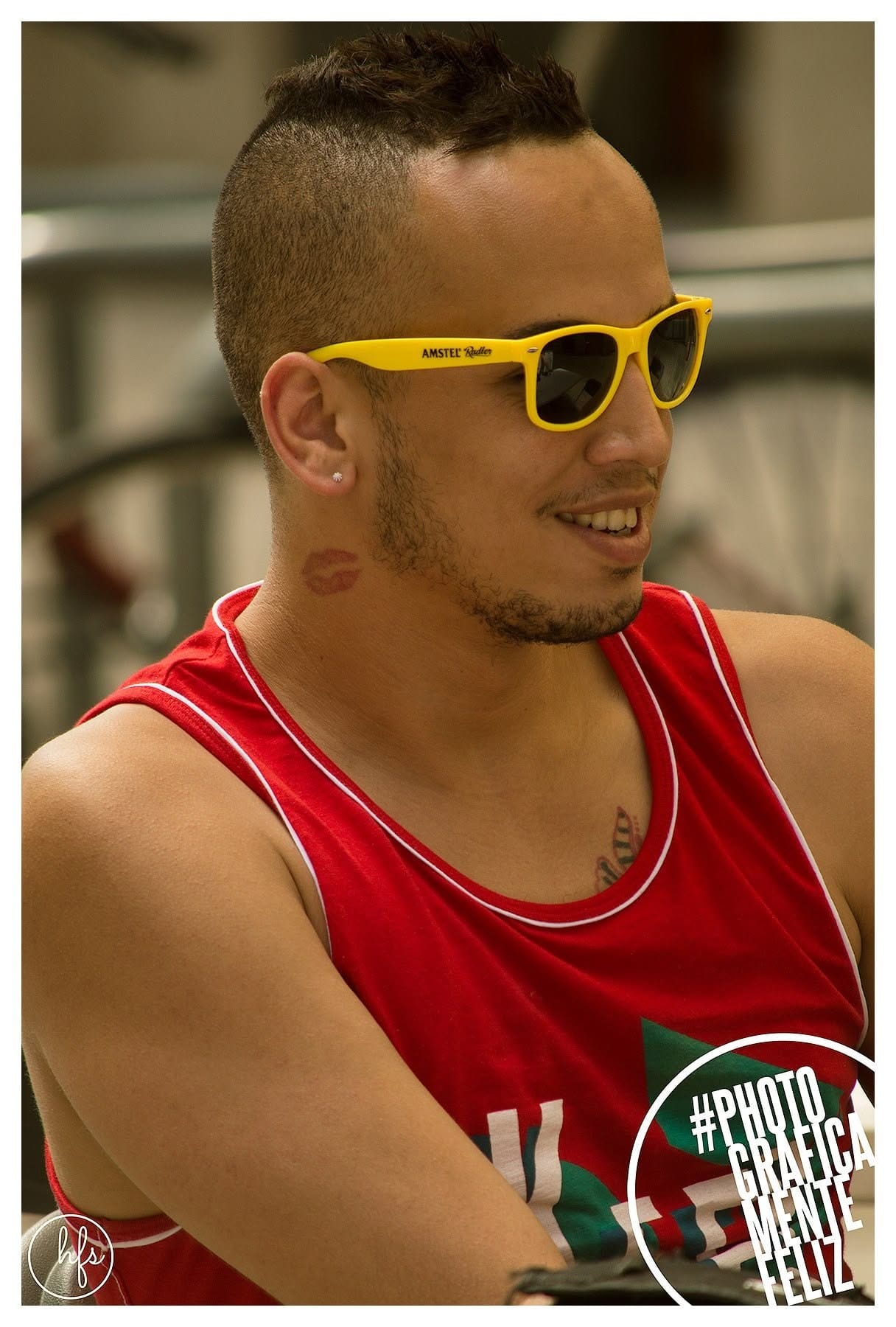 Retrato de un hombre de perfil con gafas de sol amarillas de marca de cerveza, pendiente brillante y musculosa roja, con la marca de un beso de labial dibujada en el cuello, sonriendo hacia…