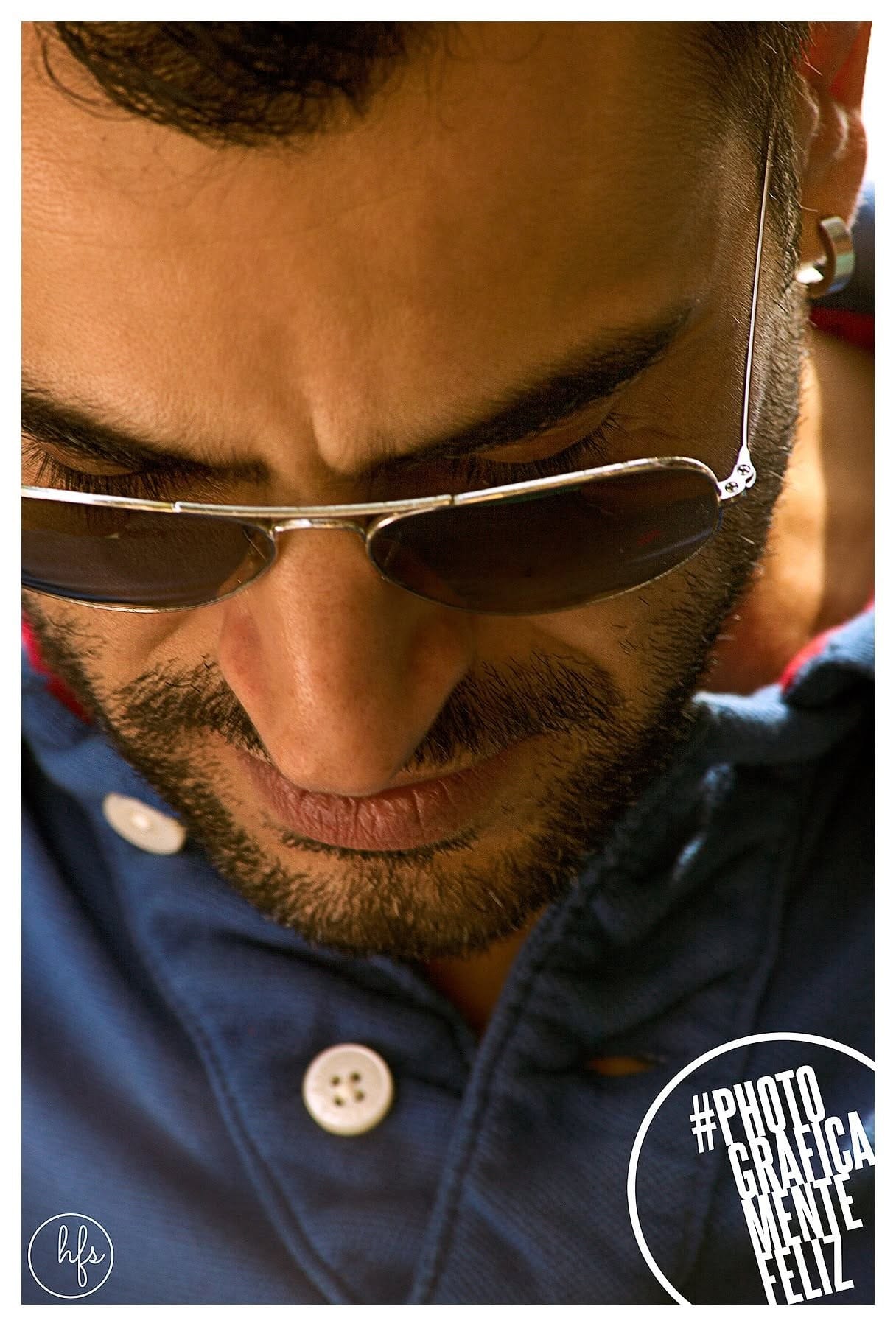 Primer plano muy cerrado de un hombre con gafas de sol de aviador mirando hacia abajo, barba corta y camisa azul con botones, iluminado por luz cálida lateral que resalta la piel y el vello…