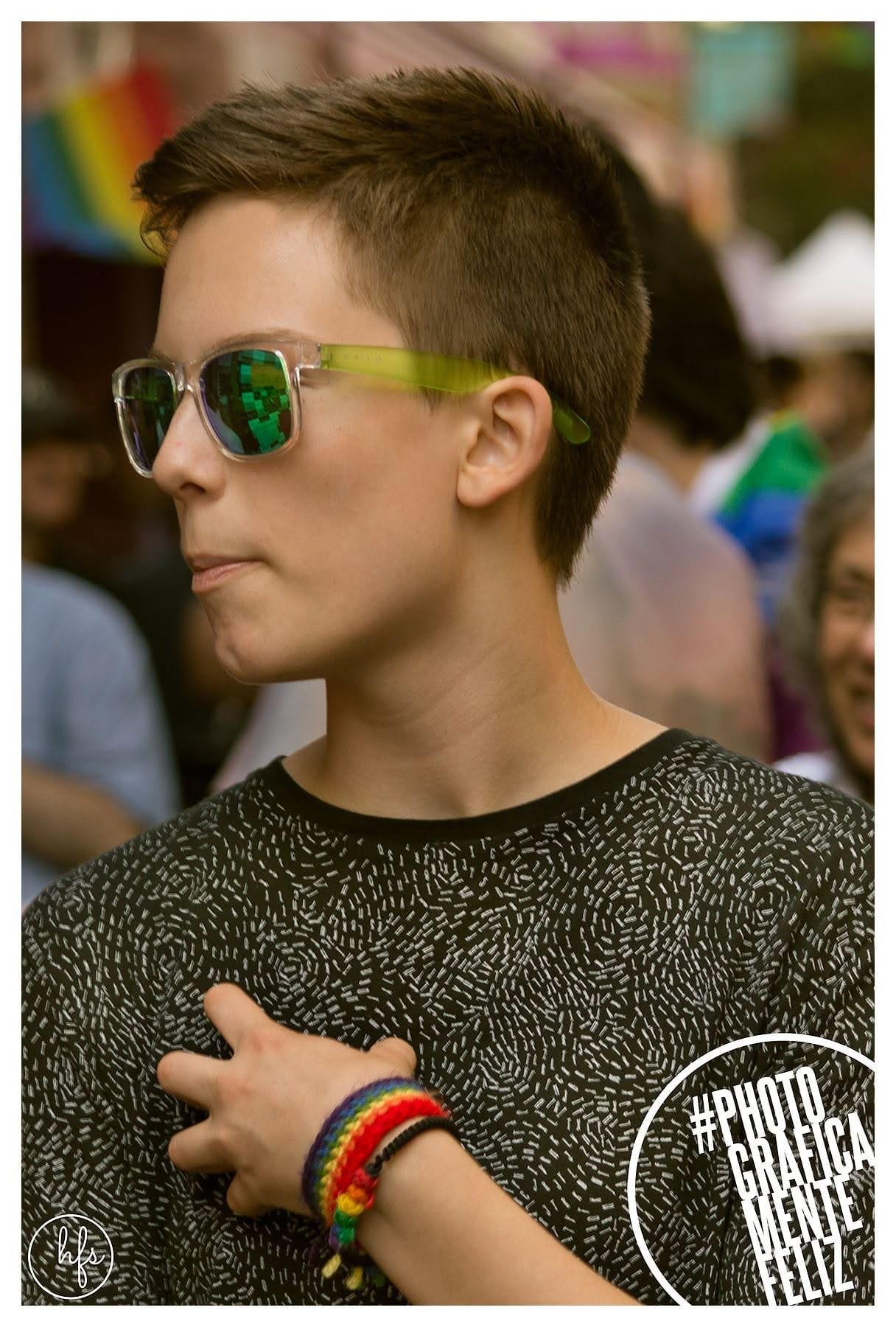 Retrato de perfil de una persona joven de pelo corto con anteojos de sol transparentes con patillas verdes, remera oscura de textura moteada, entre la multitud con una bandera del arcoíris…