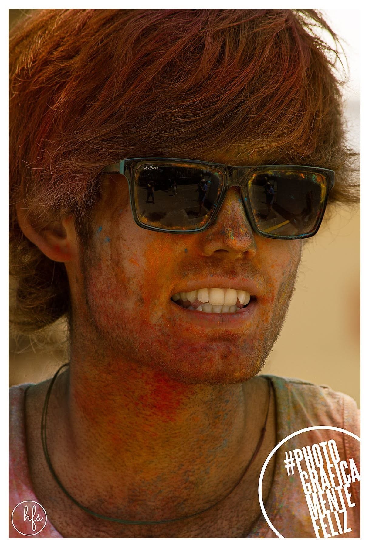 Retrato de un joven de pelo pelirrojo despeinado con gafas de sol oscuras de marca 'B-Force' que reflejan gente en la calle, cara y cuello manchados de polvo naranja, sonriendo con los…