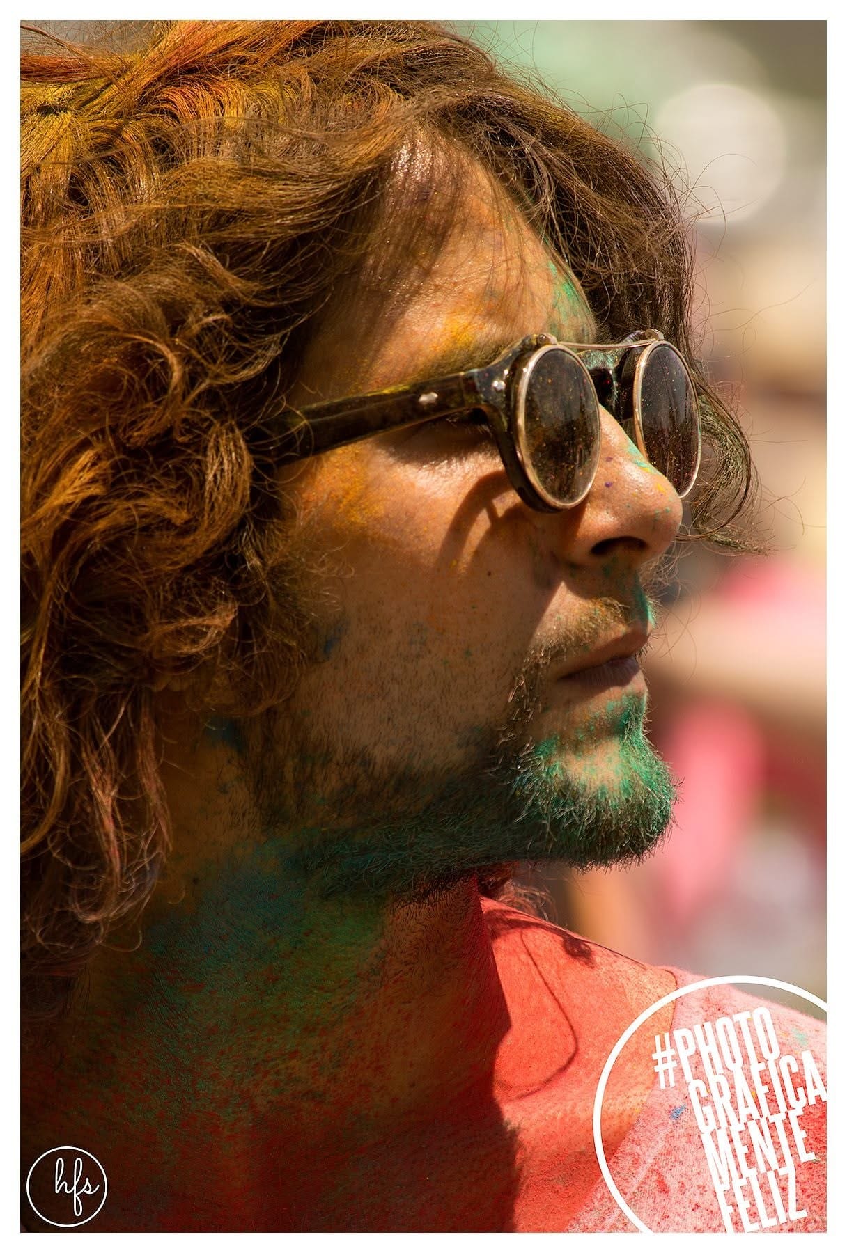 Retrato de perfil a color de un hombre de pelo rizado rojizo y gafas redondas, con la barba y parte del rostro cubiertos de polvo Holi verde y azul, mirando hacia el cielo con expresión…