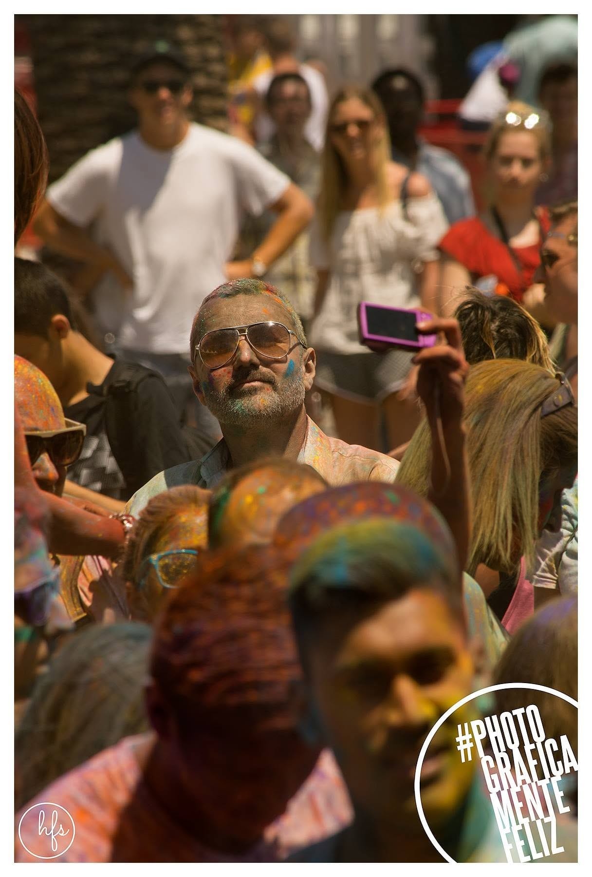 Hombre mayor de barba y pelo canos, con anteojos de sol tipo aviador, cubierto levemente de polvo de colores, fotografiando con un pequeño dispositivo en medio de una multitud durante el…