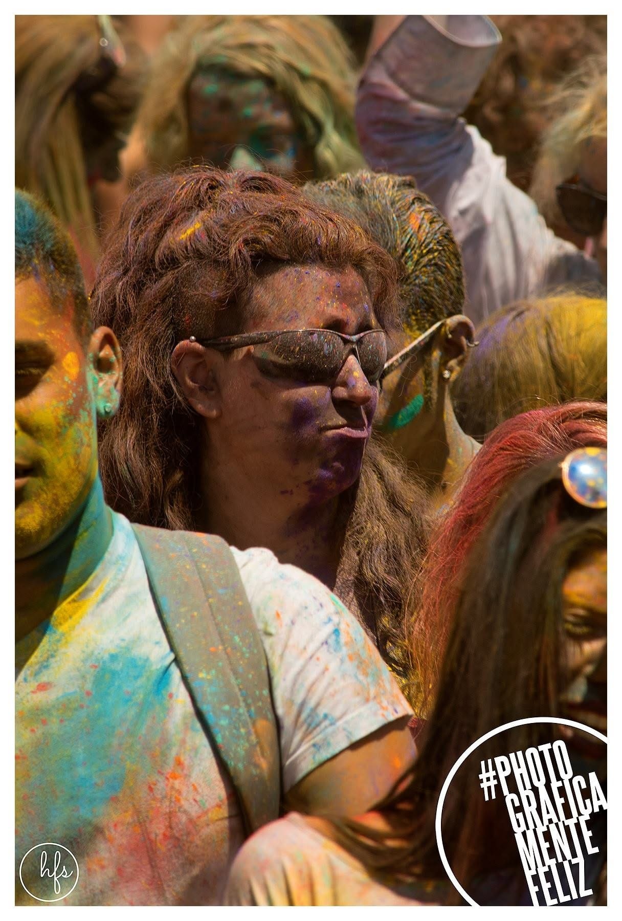 Retrato de una persona de melena castaña ondulada y anteojos de sol oscuros, rostro cubierto de pigmentos violeta y azul, rodeada muy de cerca por otras caras también pintadas de la fiesta…