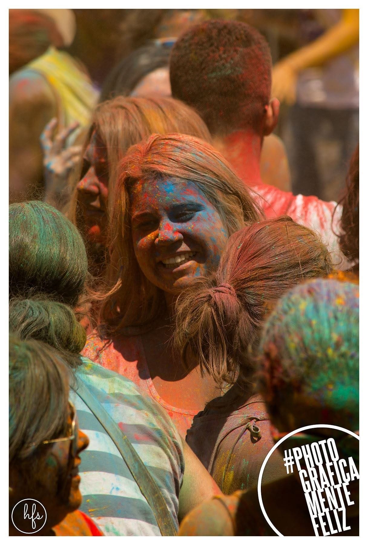 Retrato de una joven con anteojos de sol y pintura azul y roja en el rostro, sonriendo levemente, rodeada de otras personas también cubiertas de polvo de colores en un ambiente de festival.