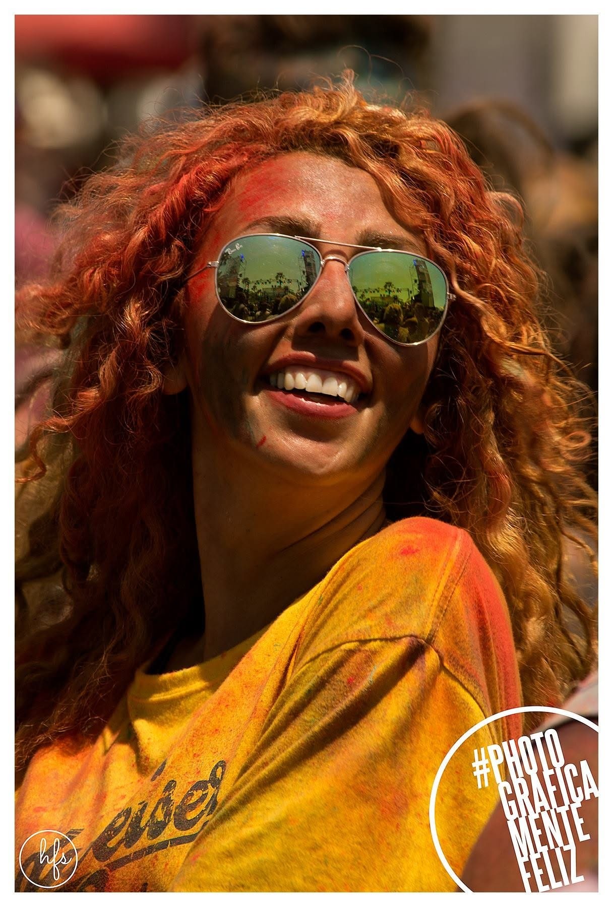 Retrato de una joven de pelo rizado pelirrojo con la piel cubierta de polvo naranja y rosa, gafas de sol espejadas que reflejan la calle, riendo con la cabeza inclinada hacia atrás bajo luz…
