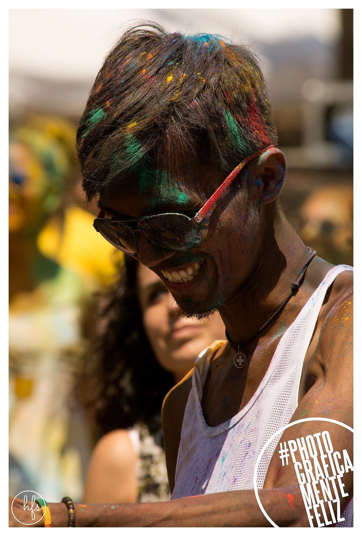 Retrato a color de un joven con el pelo y el rostro cubiertos de polvo Holi en tonos verde, amarillo y rojo, riendo con los ojos cerrados; detrás, desenfocada, una mujer también con la cara…
