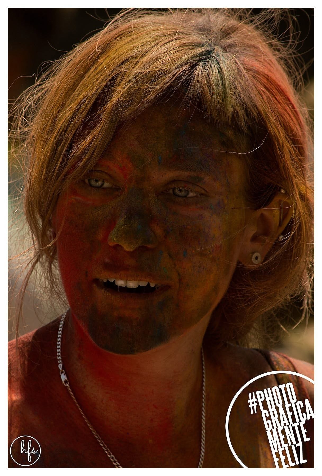 Retrato de una persona con el pelo rubio corto cubierto de tinte multicolor y la cara pintada de rojo y naranja de forma irregular, mirando fijo a cámara con expresión seria, luz cálida de…