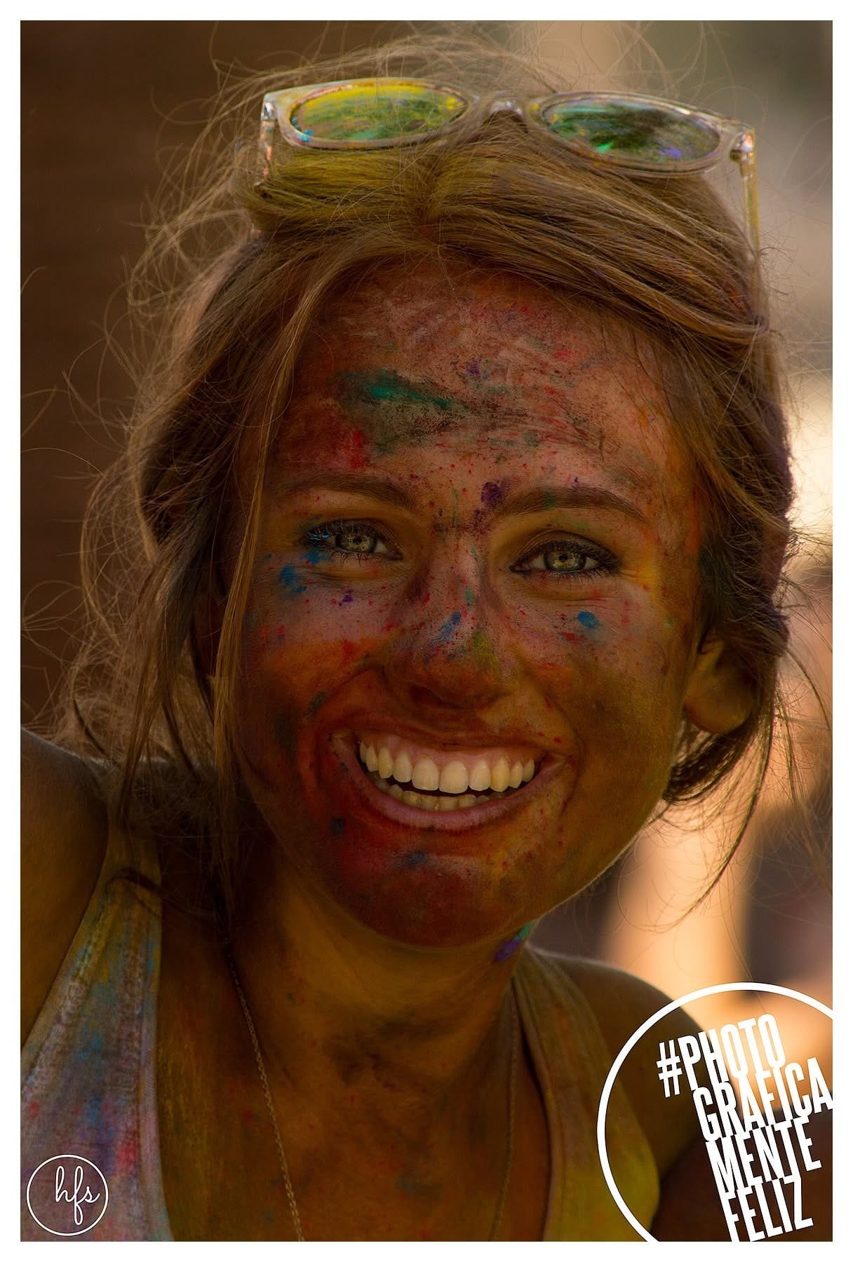 Retrato en color de una joven rubia con anteojos de sol sobre la cabeza, riendo a cámara con el rostro y el pelo cubiertos de pigmentos de colores (rojo, verde, azul, amarillo) propios de…