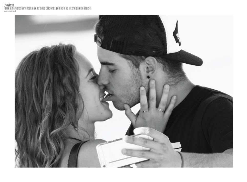 Fotografía en blanco y negro de una pareja besándose de cerca; ella con el pelo suelto sostiene el celular para la selfie, él lleva gorra plana hacia atrás.