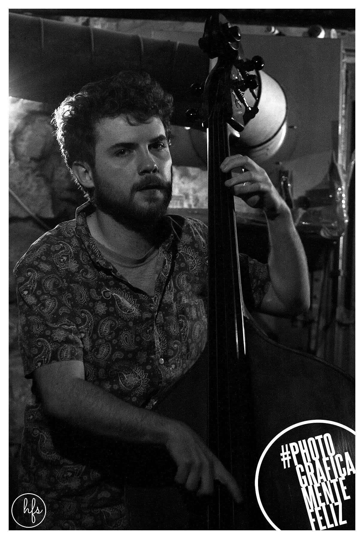 Retrato en blanco y negro de un músico de rulos oscuros y barba, con camisa estampada de cachemira, tocando un contrabajo y mirando directamente a cámara; fondo de pared de piedra con foco…