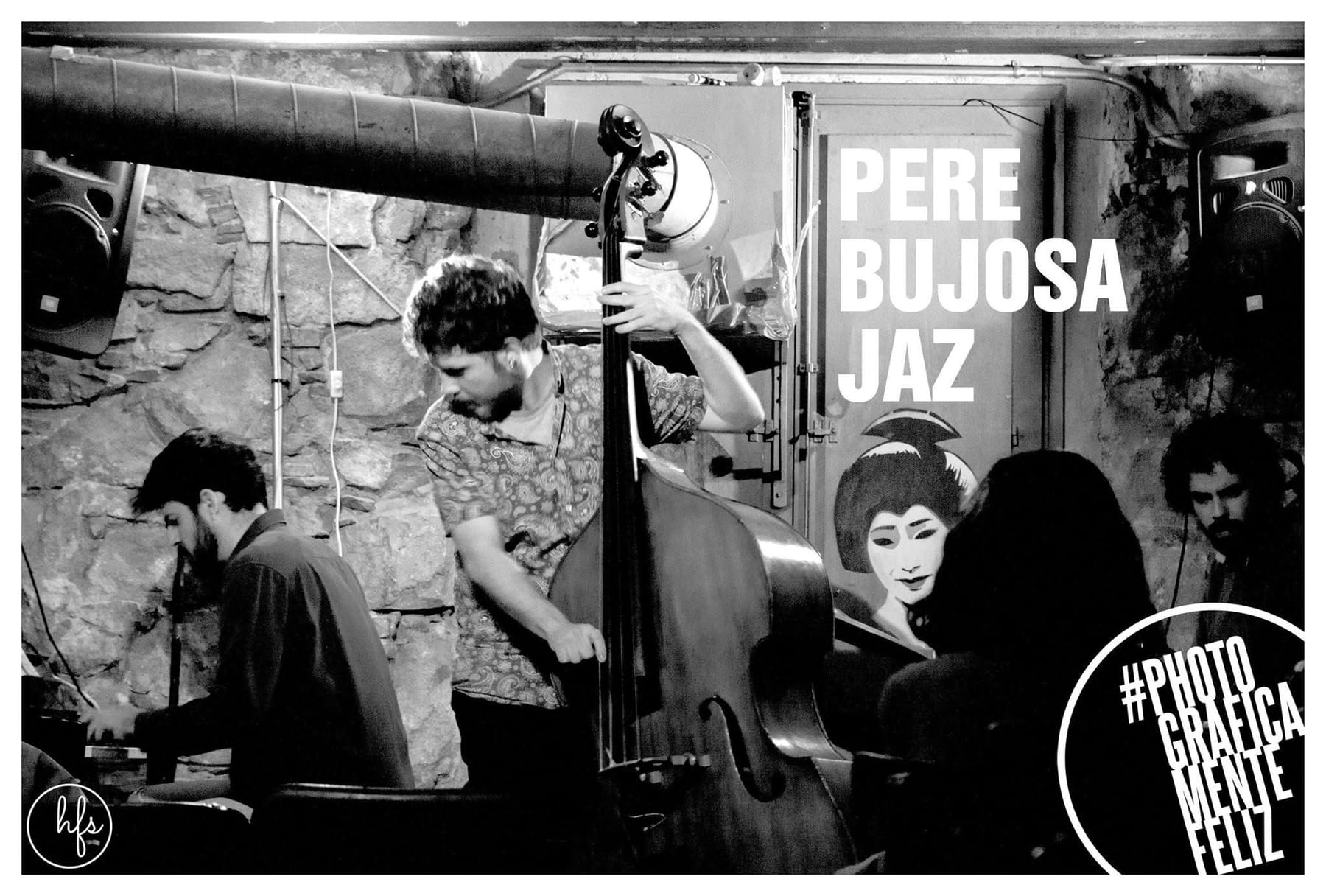 Fotografía en blanco y negro de un trío de jazz tocando en un local de paredes de piedra vista; en primer plano un contrabajista con camisa estampada apoya el instrumento junto a un cartel…