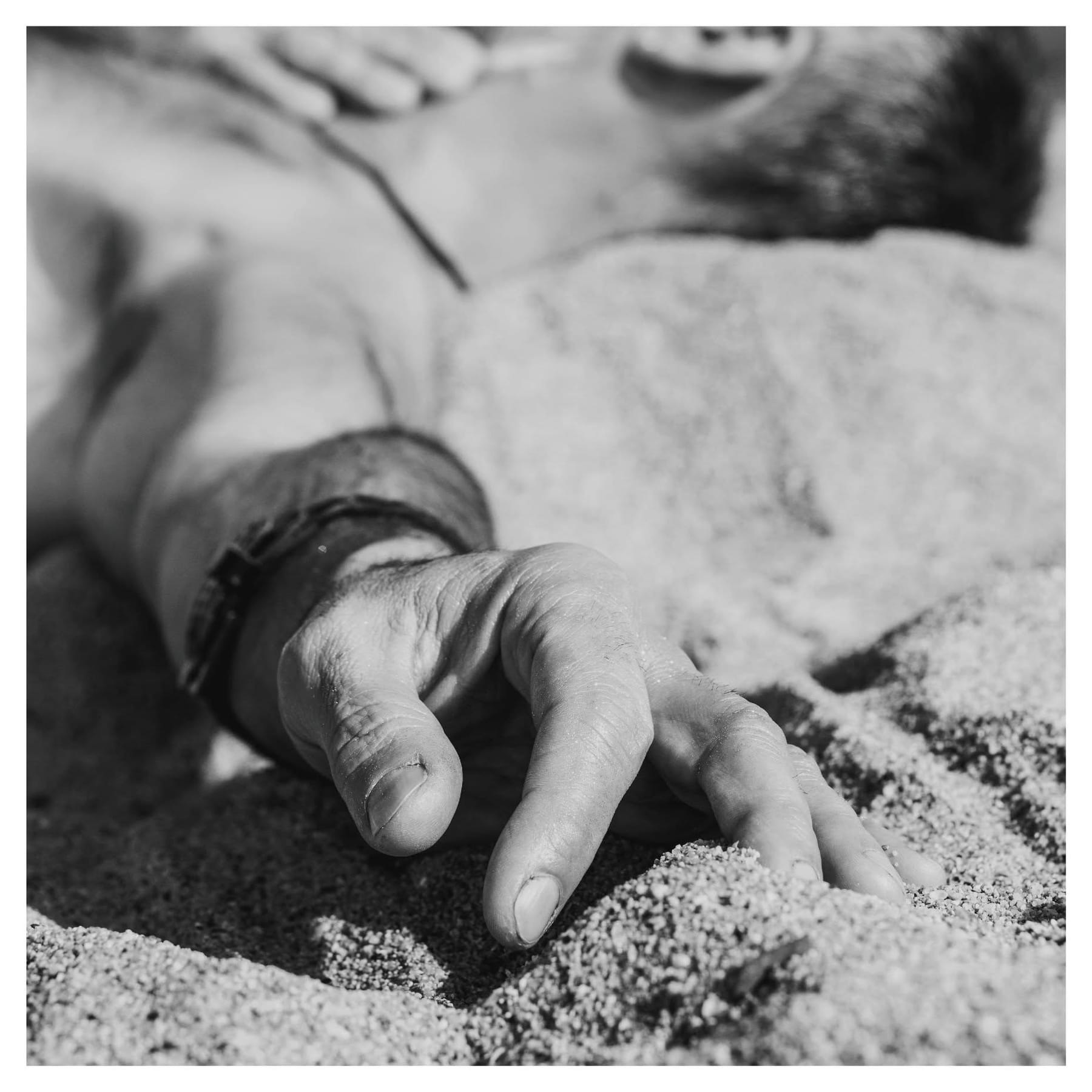 Primer plano en blanco y negro de una mano masculina apoyada sobre arena o piedra grava, con una pulsera de cuero en la muñeca; al fondo, desenfocado, se distingue un torso y otro brazo.