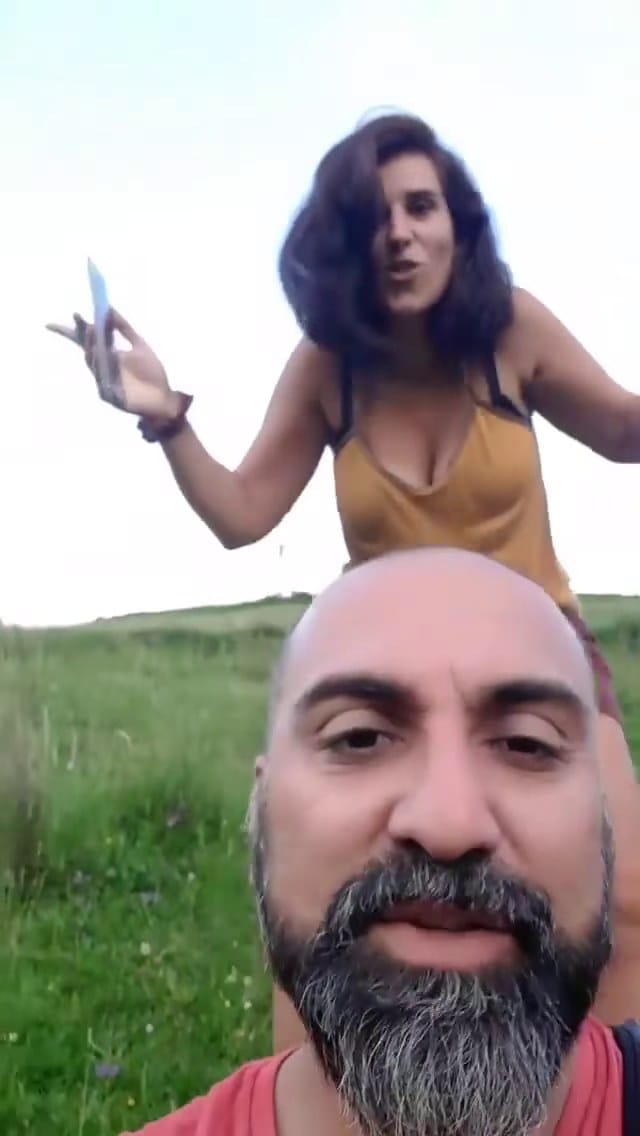 El fotógrafo graba un selfie en video en un prado de montaña, señalando hacia el cielo mientras dos personas detrás levantan los brazos también.