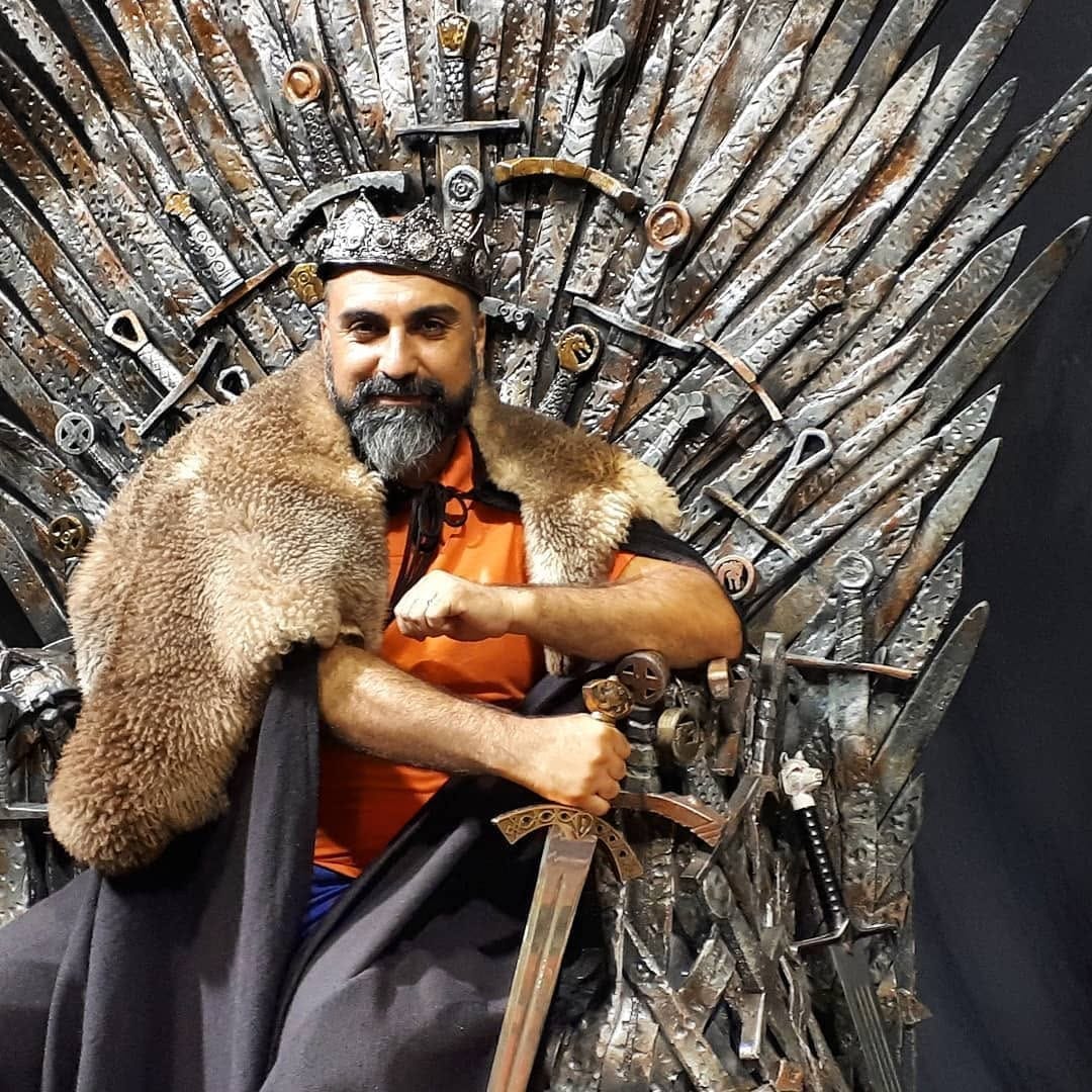 Un hombre con barba entrecana posa sentado en una réplica del Trono de Hierro de Game of Thrones, con corona, capa de piel sintética sobre los hombros y una espada en la mano.