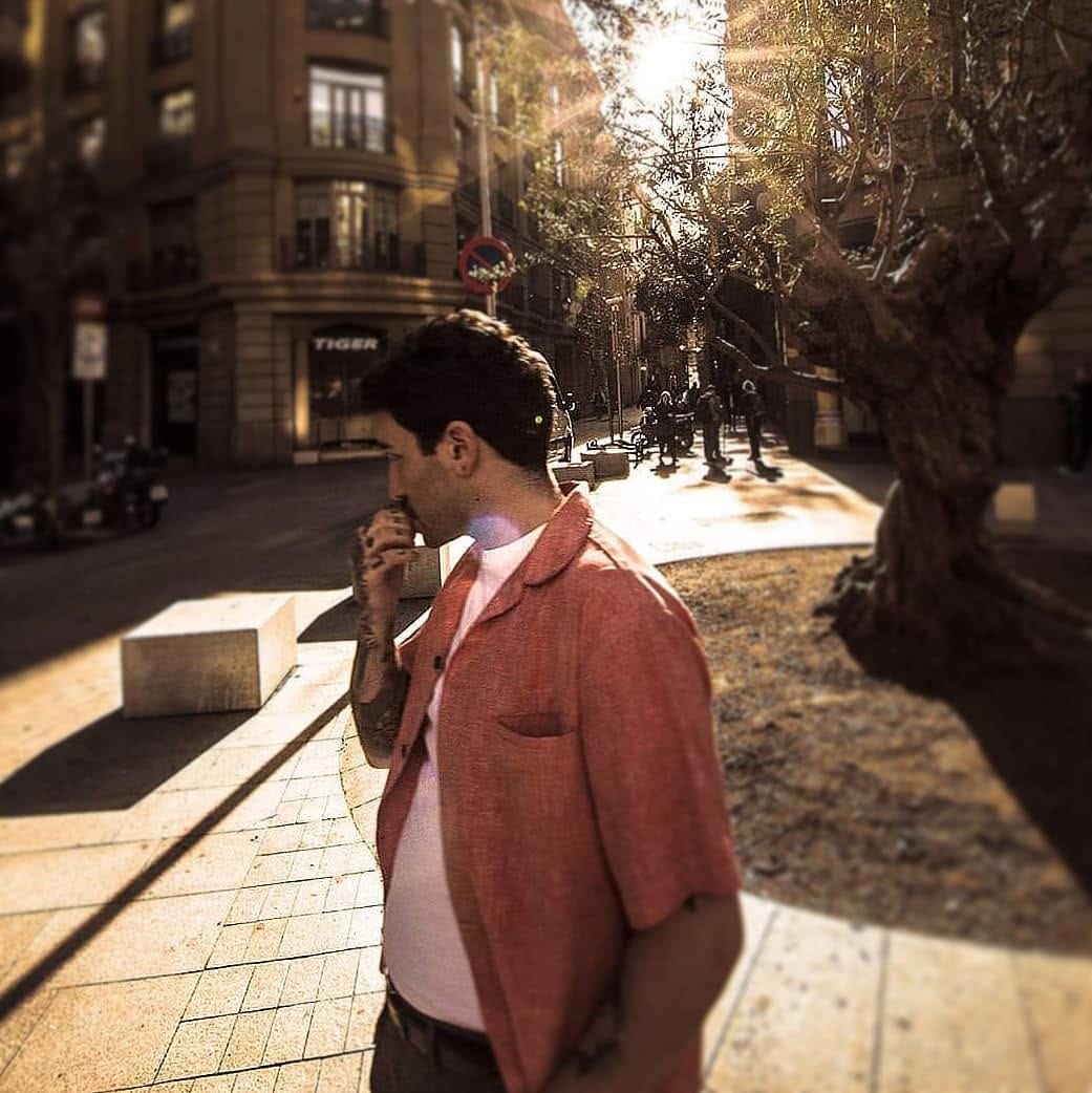 Un hombre de perfil camina por una esquina urbana arbolada, vestido con una chaqueta coral sobre remera blanca, llevándose la mano a la boca en actitud pensativa.