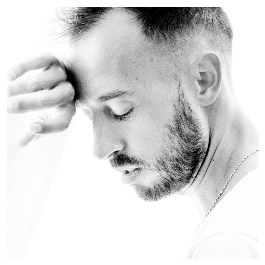 Retrato de perfil en blanco y negro de alta clave, con el modelo mirando hacia abajo y una mano apoyada en la frente en actitud reflexiva.