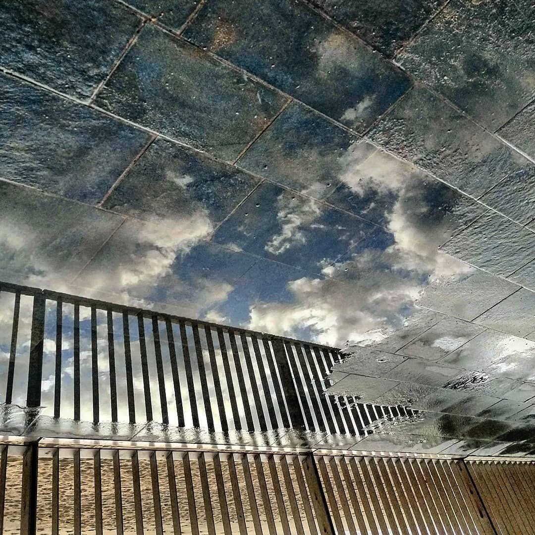 El plano esta invertido: el suelo empedrado y una baranda metalica aparecen en la mitad inferior reflejando el cielo azul con nubes blancas, mientras arriba se ve el pavimento real mojado…