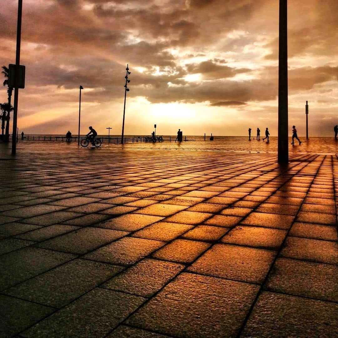 Amplia vista de un paseo empedrado junto al mar durante un amanecer o atardecer intenso, con el cielo cubierto de nubes iluminadas en tonos naranjas y dorados.