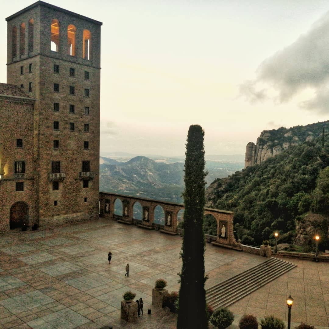 Vista amplia de la plaza y la torre del monasterio de Montserrat al atardecer, con luz calida encendida en las ventanas, un cipres alto en el centro, arcos con estatuas y la sierra de…