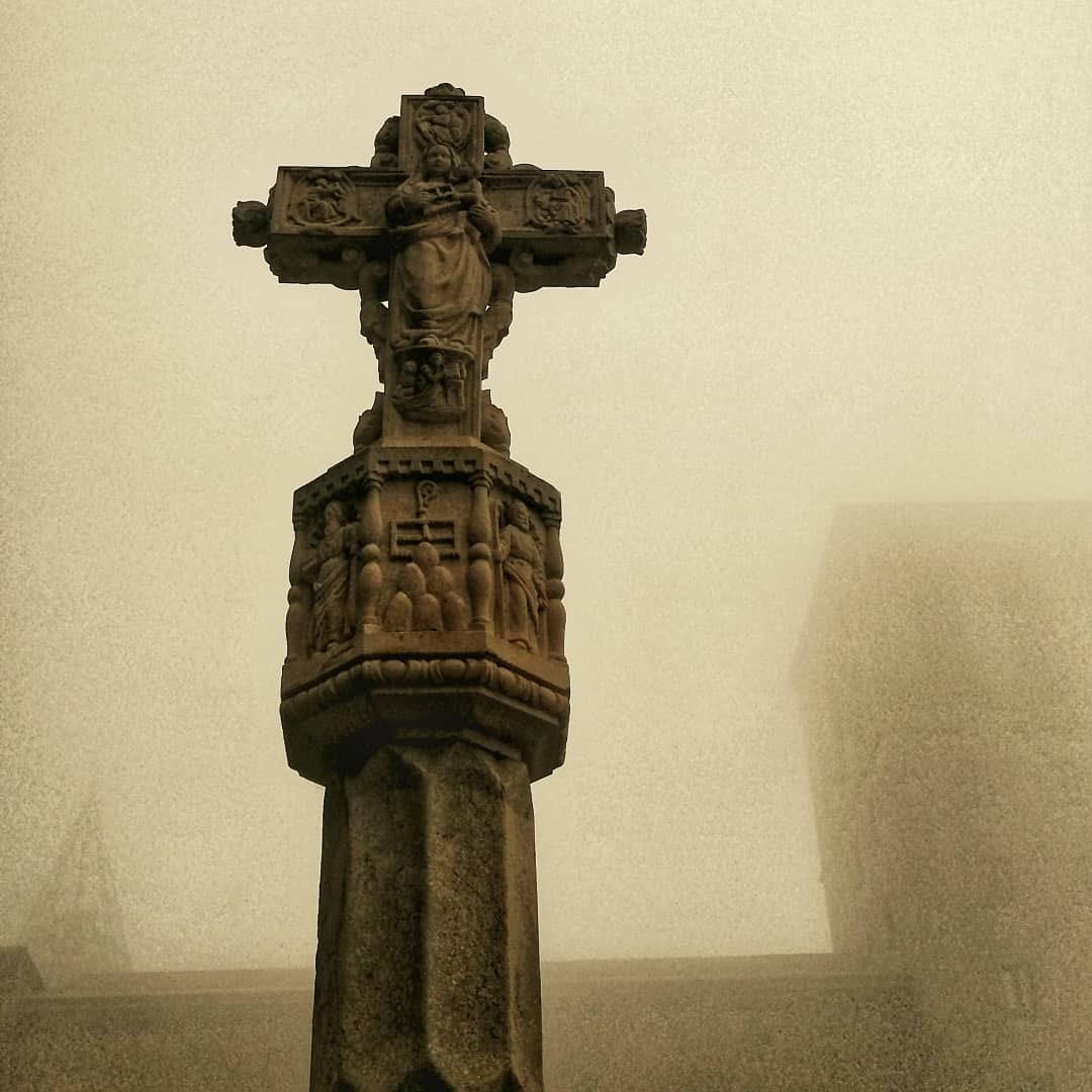 Fotografia en tono sepia de una cruz de piedra tallada con figuras religiosas sobre un pedestal ornamentado, envuelta en una niebla densa que difumina el fondo hasta casi hacerlo desaparecer.
