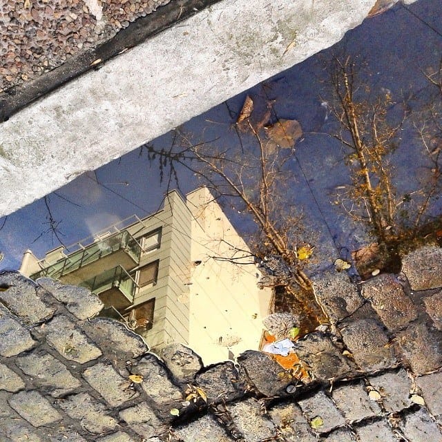 Un charco de agua sobre adoquines refleja de forma invertida un edificio de balcones, un árbol seco y un fragmento de cielo azul.