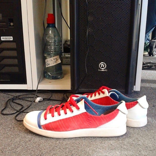 Bodegón informal en interior: un par de zapatillas rojas, blancas y azules apoyadas en el piso frente a una torre de PC negra con ventilador y una botella de agua reutilizada con etiqueta…