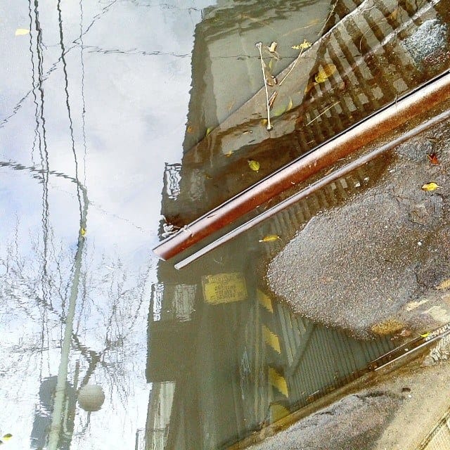 Otro charco urbano que refleja invertidos una baranda metálica oxidada, la fachada de un edificio y hojas caídas sobre el pavimento.