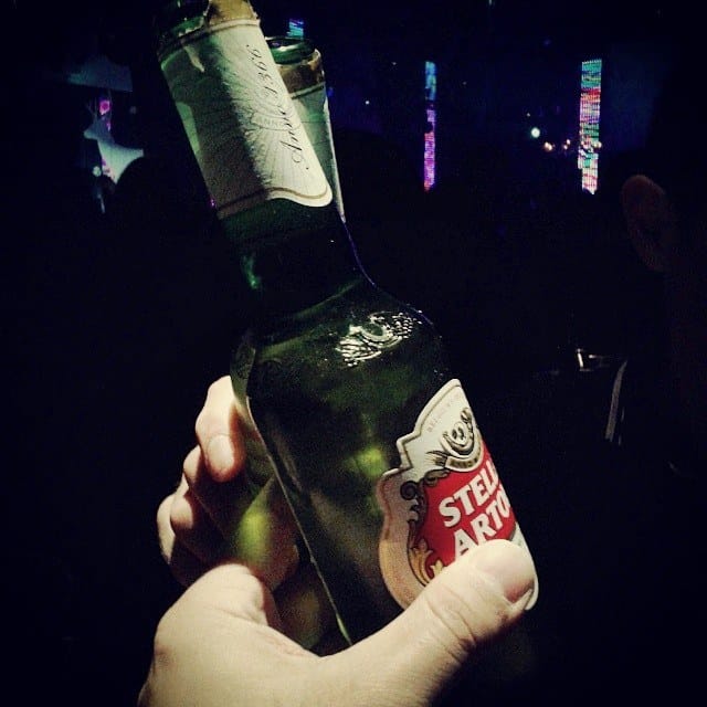Primer plano de una mano sosteniendo una botella de cerveza Stella Artois, con luces de discoteca desenfocadas al fondo en tonos violeta y turquesa.