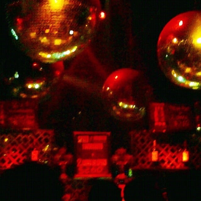 Composición casi abstracta de esferas decorativas doradas y rojas colgando sobre una barra iluminada en tonos rojo intenso, con botellas y una reja de madera al fondo.