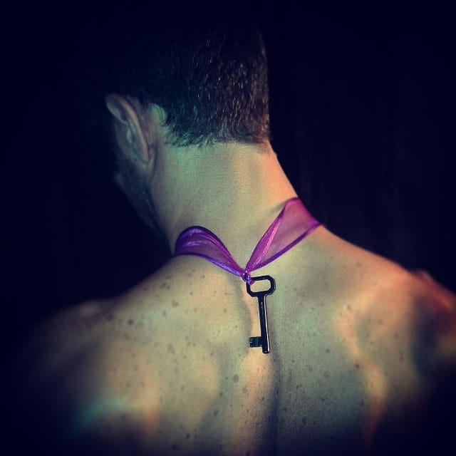 Primer plano de la espalda desnuda de un hombre joven, con el pelo corto oscuro, del que cuelga una llave antigua atada con una cinta de organza violeta al cuello.