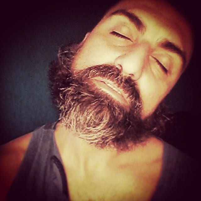 Retrato cercano de un hombre calvo con barba tupida y ojos cerrados, en musculosa gris, iluminado de forma suave y cálida sobre fondo oscuro azulado.