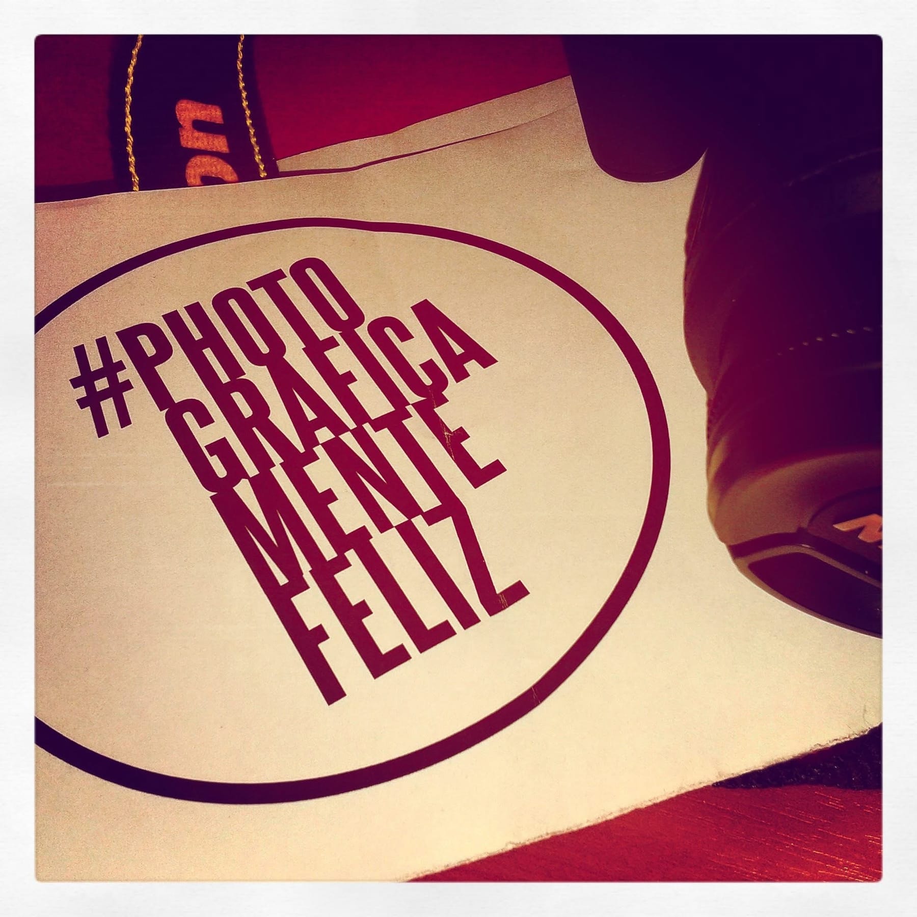 Fotografía de una tarjeta impresa con el logo "#photograficamentefeliz" en un óvalo bordó sobre papel claro, apoyada junto al borde de una zapatilla oscura sobre un piso rojizo.