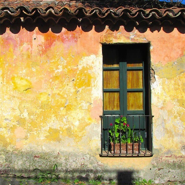 Fachada colonial de pared ocre y rosada desgastada por el tiempo, con un balcón de rejas negras, puertas de madera verde oscuro y macetas con flores rojas y rosas.