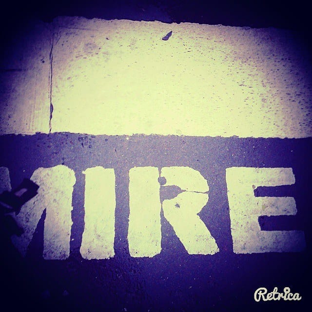 Detalle a nivel del piso de la palabra "MIRE" pintada con plantilla blanca sobre asfalto oscuro, junto al borde de una vereda de cemento claro.