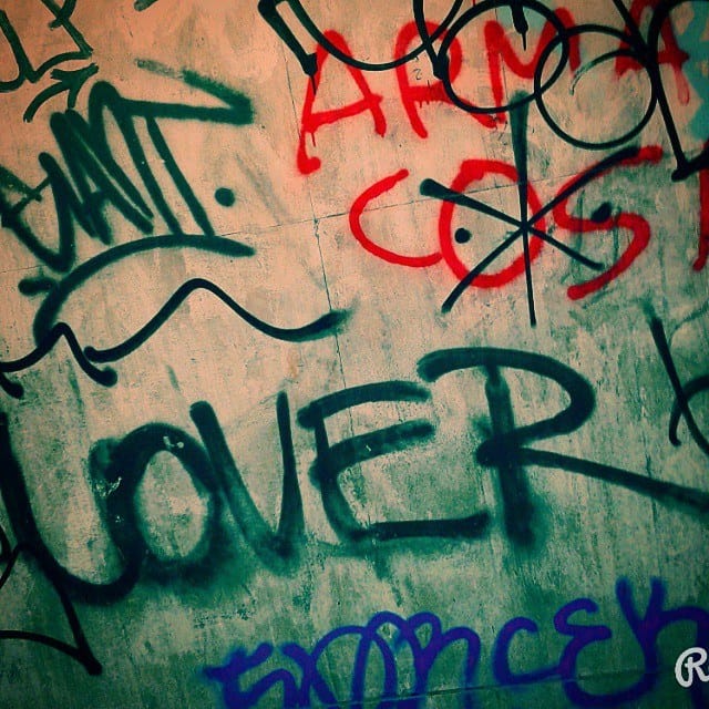 Pared cubierta de grafitis superpuestos en verde, rojo, negro y azul, donde destaca la palabra "LOVER" en letras verdes de gran tamaño rodeada de tags y firmas menores.